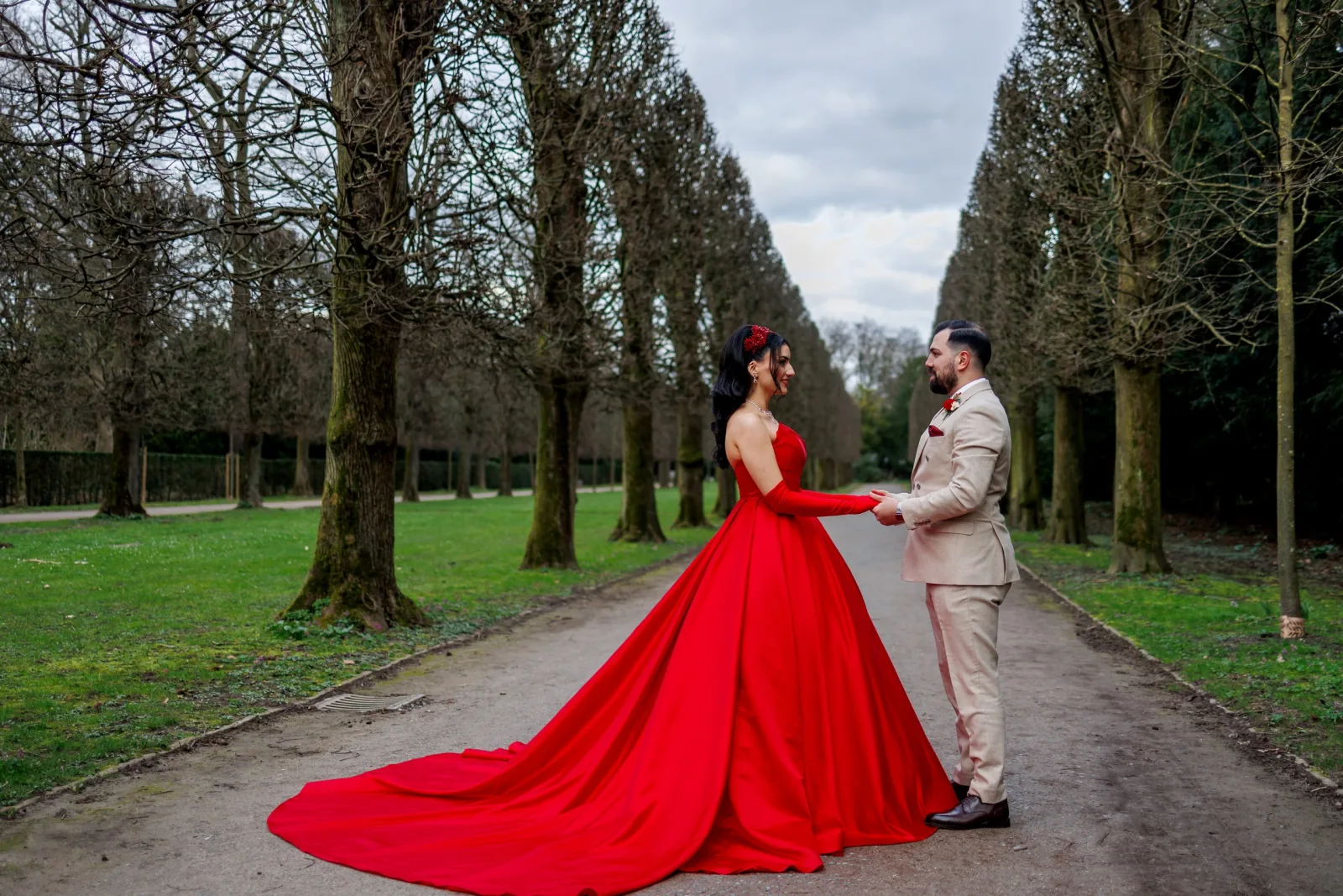 Couple holding hands in a tree-lined alley, woman in a red gown